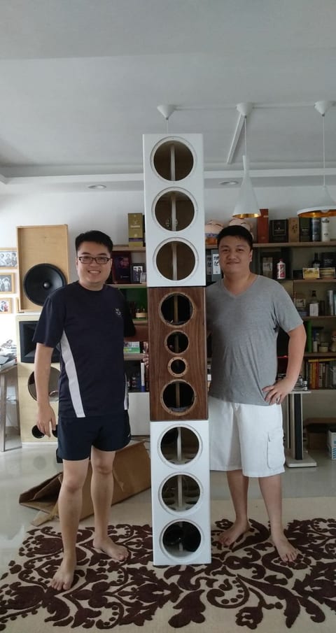 Two men stand either side of a tall stack of three speaker cabinet sections with open circular driver cut-outs.