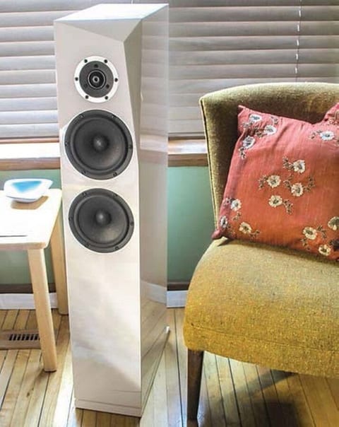 Slim white floorstanding tower speaker with a tweeter and two cone drivers, next to an armchair with a cushion.