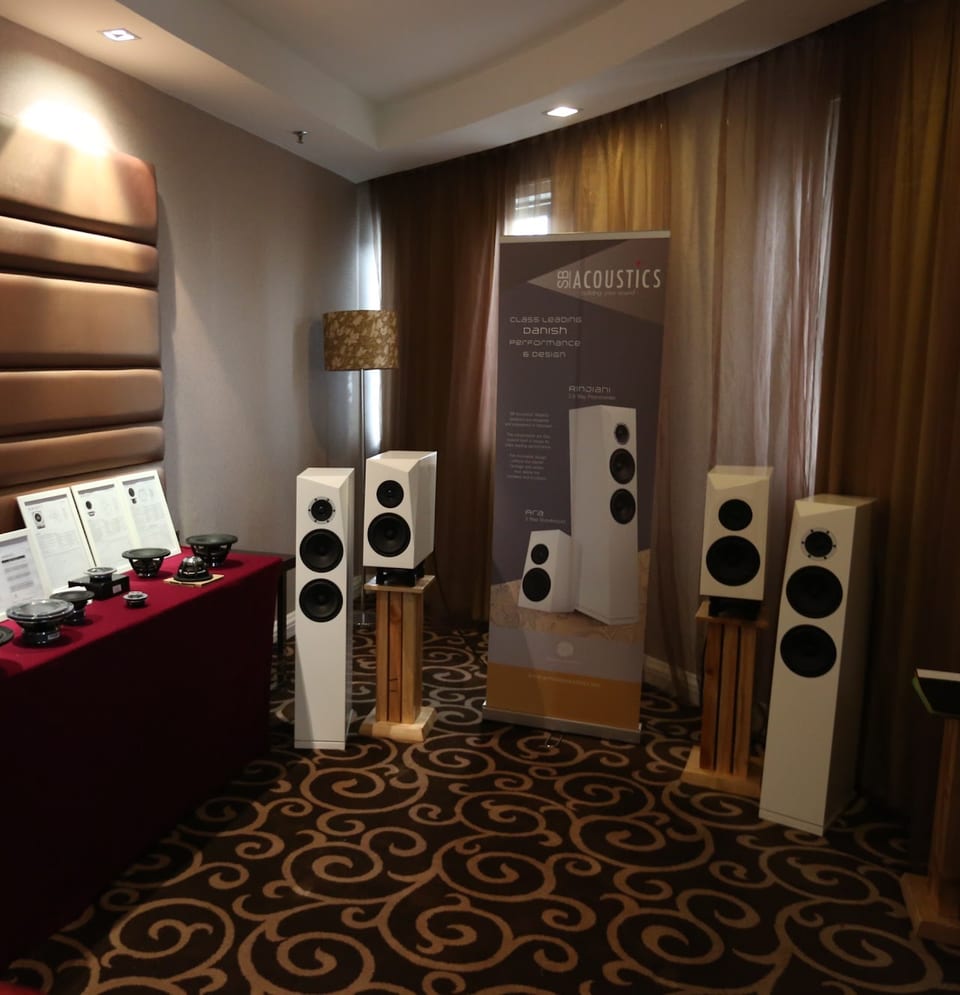 White SB Acoustics floorstanders and standmounts in a demo room, with an SB Acoustics banner and drivers on a table