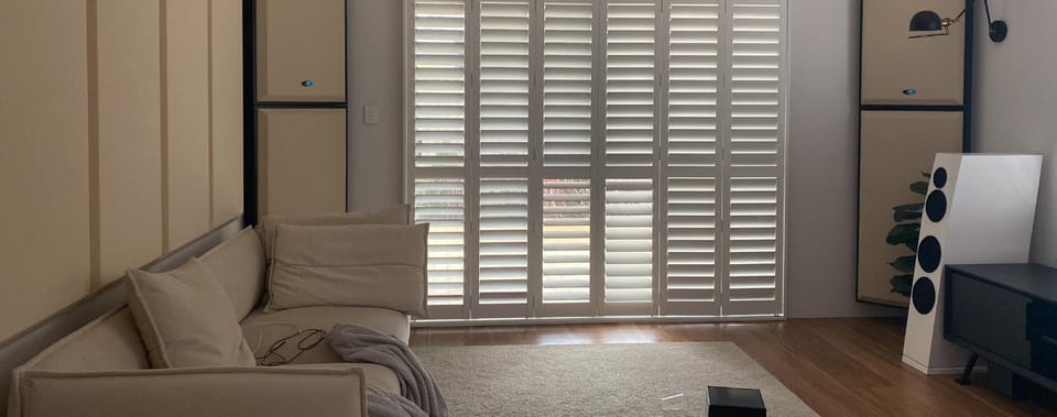 Living room with a beige sofa, plantation shutters, and a white four-driver floorstanding speaker at right