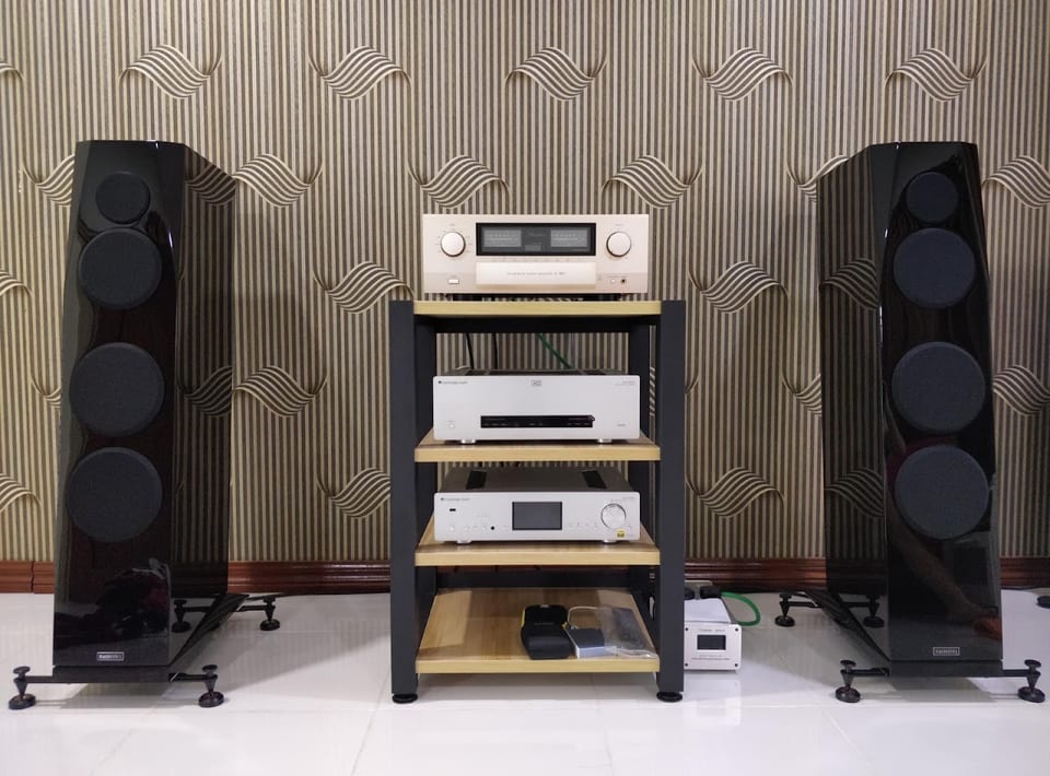 Pair of gloss black Sasandu floorstanders on outrigger feet either side of a four-shelf rack of hi-fi electronics.