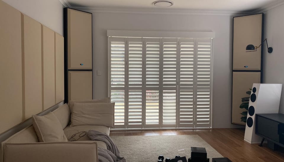 Living room with plantation shutters and beige sofa; white SB Acoustics Sasandu floorstander at right.